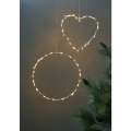 Ciepłe dekoracje LED w formie serca i koła, wiszące na ciemnej ścianie obok gałązek sosny.