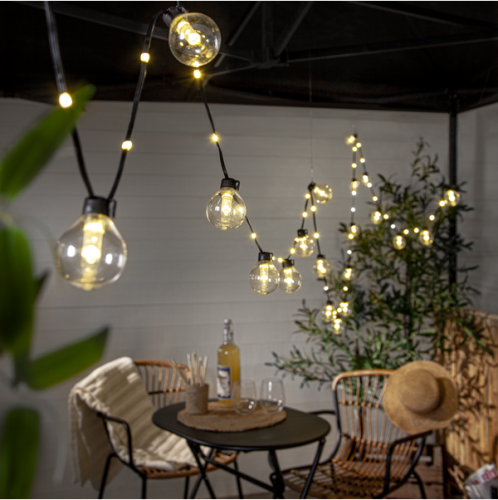 Girlanda LED Bolla z przezroczystymi kulami emitującymi ciepłe białe światło, zawieszona nad stołem na tarasie, tworząca przytulną atmosferę wieczornego wypoczynku.