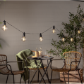 Girlanda ogrodowa LED Circus Nevada z przezroczystymi kloszami i żarówkami filament, zawieszona nad stolikiem na tarasie, ciepłe białe światło tworzące przytulną atmosferę.