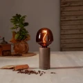 Dekoracyjna żarówka LED filament z bursztynowego szkła ustawiona na lampie stołowej na drewnianym blacie obok rozsypanych ziaren kawy. Designerskie oświetlenie emitujące ciepłe światło.