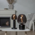 Dwie dekoracyjne żarówki LED z przydymionego szkła ustawione na czarnych lampach stołowych na białym stoliku przed kominkiem. Designerskie oświetlenie emitujące ciepłe światło.