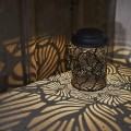 Solarny lampion LED z czarną ażurową obudową tworzący dekoracyjne cienie na blacie i drewnianej ścianie.