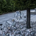 Dwie solarne kule dekoracyjne z metalowych obręczy umieszczone w kamiennej aranżacji ogrodowej. Jedna z ozdób zawieszona jest na gałęzi, a delikatne światło LED podkreśla przestrzenną formę obu dekoracji.