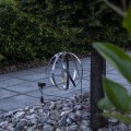 Wisząca dekoracja solarna w formie metalowej kuli zamocowana na gałęzi nad kamienną rabatą. Ażurowa konstrukcja z przecinających się obręczy została podświetlona niewielkim źródłem światła LED.