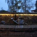 Wąż świetlny LED zamontowany wzdłuż ceglanej ściany w ogrodzie, delikatnie podświetlający otoczenie z metalową konewką i rośliną w tle.