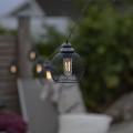 Zbliżenie metalowego lampionu LED z girlandy solarnej. Czarna, siateczkowa obudowa oraz widoczna żarówka LED w stylu Edison emitująca ciepłe światło.