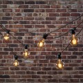 Girlanda ogrodowa MIMOSA z żarówkami G80 zawieszona przed ceglaną ścianą. Ciepłe światło LED 2000K, czarny przewód i dekoracyjny układ linii świetlnej.