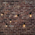 Girlanda ogrodowa MIMOSA z żarówkami G45 zawieszona przed ceglaną ścianą. Ciepłe światło LED 2100K, czarny przewód i dekoracyjny układ linii świetlnej.
