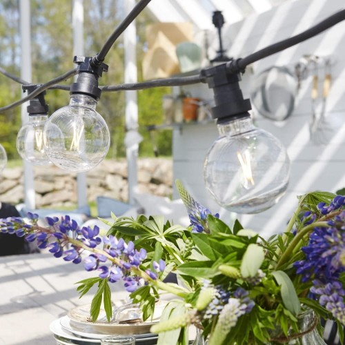 Girlanda ogrodowa z dużymi, przezroczystymi plastikowymi żarówkami G95 LED 1W 2700K, zawieszona nad stołem z bukietem fioletowych kwiatów w jasnej altanie w słoneczny dzień.