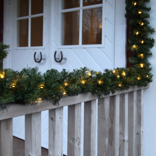 Zielona girlanda z ciepłymi lampkami LED ułożona na drewnianej balustradzie przed białymi drzwiami z przeszkleniami.