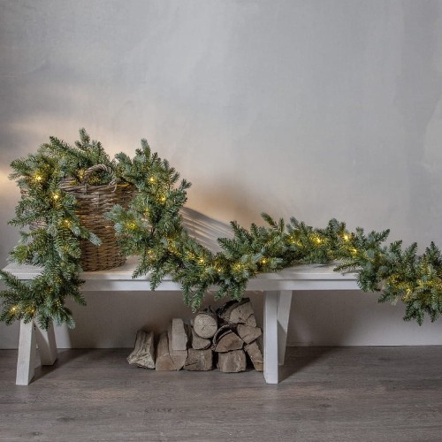 Zielona girlanda świerkowa LED zwisająca z wiklinowego kosza ustawionego na białej ławce, z długimi gałązkami opadającymi na podłogę i ciepłym światłem tworzącym świąteczną atmosferę.