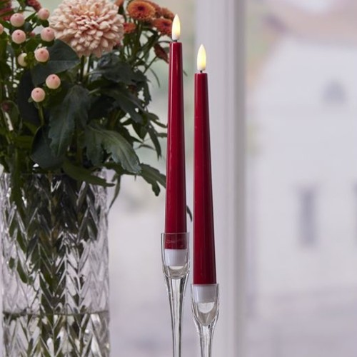 Czerwone smukłe świece LED w szklanych świecznikach obok wazonu z kwiatami na stole przy oknie.
