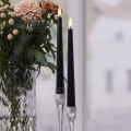 Dwie smukłe czarne świece LED w szklanych świecznikach obok wazonu z kwiatami na stole przy oknie.