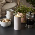 Beżowa świeca LED w kształcie walca na stole obok filiżanek, miski z ciasteczkami, kawiarki i doniczkowej rośliny.