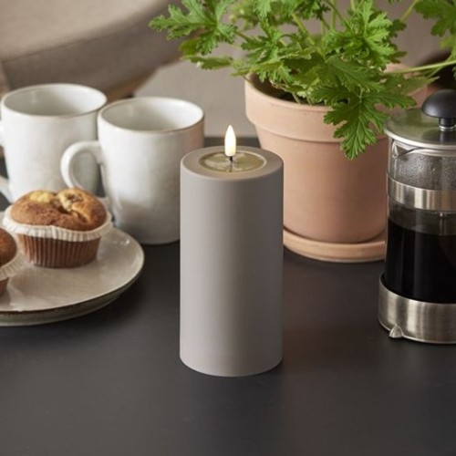 Szara świeca LED w cylindrycznym kształcie na stole obok filiżanek, muffinek i kawy w zaparzaczu French press, w tle doniczka z zieloną rośliną.