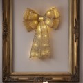 Złota dekoracja LED w kształcie kokardy zawieszona w ozdobnej złotej ramie na jasnej ścianie, tworząca elegancki wystrój wnętrza.