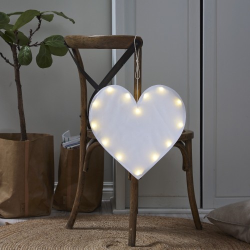 Biała dekoracja LED w kształcie serca, podświetlana ciepłym światłem, zawieszona na oparciu drewnianego krzesła w przytulnym wnętrzu.