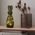 Szklana figurka zająca LED w odcieniu zieleni na drewnianej półce; w środku ciepłe, delikatne światło, obok ceramiczny wazon z suchymi roślinami. Spokojna, nastrojowa dekoracja do wnętrza.