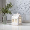 Biały podświetlany domek LED wypełniony brokatowym płynem wirującym jak śnieg, stojący na białym parapecie obok szklanej butelki z gałązką, na tle zimowego okna.