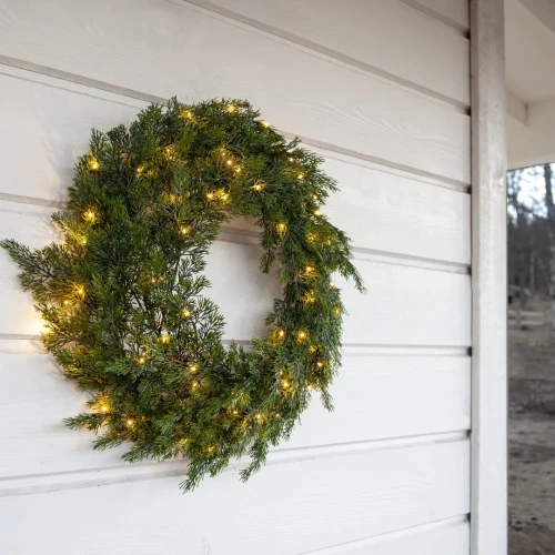 Zielony wieniec z drobnymi światełkami LED zawieszony na jasnej ścianie domu, wprowadzający ciepły, świąteczny nastrój.