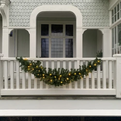 Zielona girlanda z gęstymi iglastymi gałązkami i ciepłymi lampkami LED dekorująca białą balustradę werandy, z dużym oknem w tle.