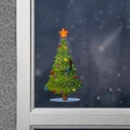 Świąteczna naklejka LED w kształcie choinki z kolorowymi ozdobami i świecącą gwiazdą, umieszczona na oknie z widokiem na ciemne, zimowe tło.