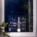 Naklejka LED z rysunkiem zimowego miasteczka i świecącą gwiazdą na szczycie jednego z domów, umieszczona na okiennej szybie z nocnym, zimowym tłem i gałązkami świerku po bokach.