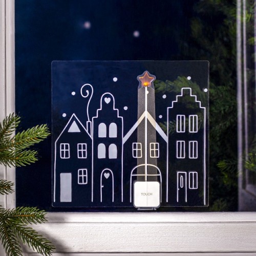 Świąteczna naklejka LED przedstawiająca rysunek zimowego miasteczka z podświetloną gwiazdą na szczycie jednego z domów, umieszczona na szybie okna z ciemnym, zimowym tłem i gałązkami świerku.