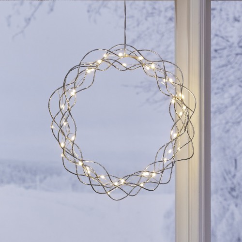 Srebrny wianek LED wykonany z cienkich, przeplatających się drucików, podświetlony ciepłymi punktami światła i zawieszony w oknie z widokiem na ośnieżony krajobraz.
