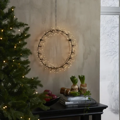 Czarna dekoracja LED w formie okręgu z drobnymi, gwiazdkowymi punktami ciepłego światła, zawieszona na ścianie obok choinki i stołu udekorowanego roślinami, z zimowym widokiem za oknem.