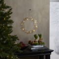 Złoty wianuszek dekoracyjny LED z delikatnymi listkami i drobnymi gwiazdkami w ciepłym świetle, zawieszony na ścianie obok choinki i dekoracyjnych roślin, z zimowym widokiem za oknem.