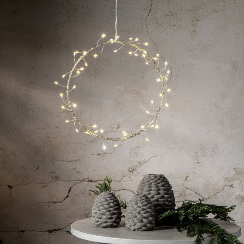 Wianek LED na subtelnej, metalowej ramie zawieszony na tle jasnej, lekko postarzonej ściany, roztaczający świąteczny blask nad stołem ozdobionym szyszkami i zielonymi gałązkami.