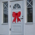 Czerwona dekoracja LED w kształcie dużej świątecznej kokardy, zawieszona na białych drzwiach wejściowych domu.