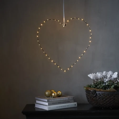 Delikatne serce LED zawieszone na jasnej wstążce, subtelnie rozświetlające wnętrze ciepłym blaskiem. Dekoracja tworzy przytulny nastrój nad stolikiem z książkami i złotymi akcentami.