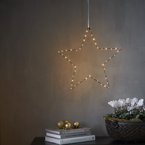 Subtelna gwiazda LED zawieszona na jasnej wstążce, roztaczająca ciepłe, przytulne światło nad stolikiem z książkami i złotymi ozdobami. Delikatna dekoracja, która ociepla wnętrze miękkim blaskiem.