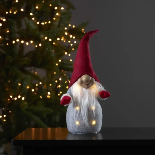 Dekoracyjny skrzat LED z długą białą brodą i czerwoną czapką stojący na czarnym blacie, w tle rozświetlona choinka.