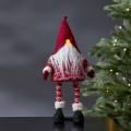 Świąteczna figurka skrzata LED w czerwonej czapce i sweterku, z długą białą brodą i świecącym noskiem, stojąca na czarnym blacie obok choinki z ciepłymi lampkami.