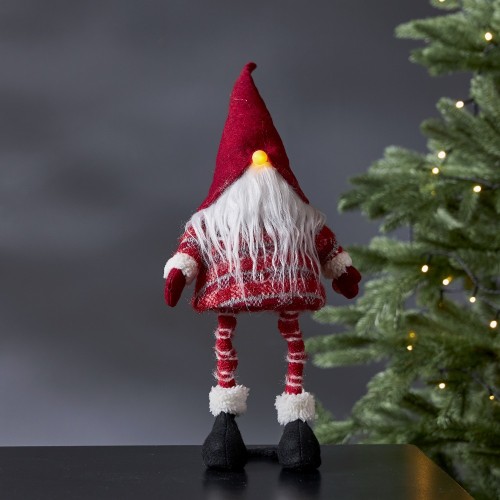 Świąteczna figurka skrzata LED w czerwonej czapce i sweterku, z długą białą brodą i świecącym noskiem, stojąca na czarnym blacie obok choinki z ciepłymi lampkami.