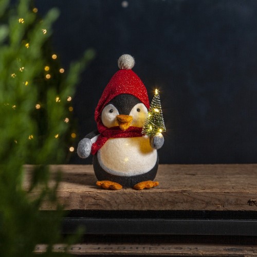 Świąteczna figurka pingwina LED w czerwonej czapce i szaliku, trzymającego małą podświetlaną choinkę, stojącego na drewnianym blacie w przytulnej, zimowej scenerii.