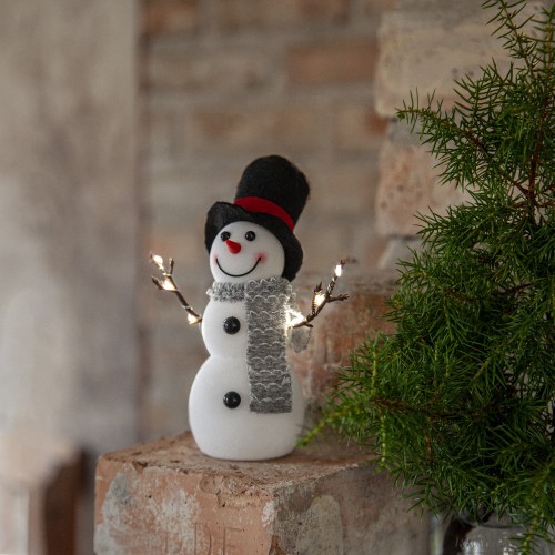 Świąteczna figurka bałwanka LED w cylindrze i szarym szaliku, z delikatnie podświetlanymi gałązkami, stojąca na ceglanej półce obok zielonej rośliny, tworząca przytulny zimowy nastrój.