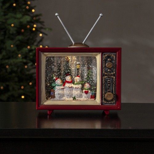 Podświetlany świąteczny telewizor retro LED w czerwonej obudowie, w środku scena z rodziną bałwanków na tle choinek w wirującym brokacie, ustawiony na ciemnym blacie przy choince z lampkami.