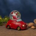 Świąteczna figurka z Mikołajem w śnieżnej kuli, osadzona w czerwonym autku z subtelnie podświetlanymi reflektorami, ustawiona na drewnianym blacie obok gałązki z lampkami i orzechów. Przytulna, zimowa aranżacja wnętrza.
