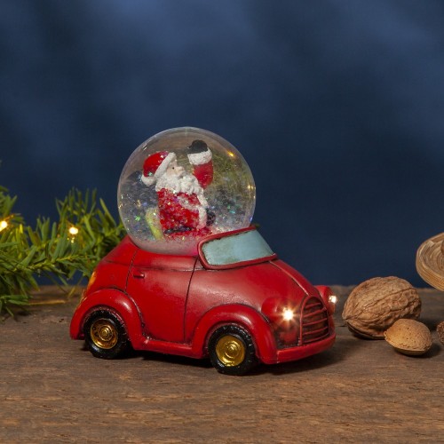 Świąteczna figurka z Mikołajem w śnieżnej kuli, osadzona w czerwonym autku z subtelnie podświetlanymi reflektorami, ustawiona na drewnianym blacie obok gałązki z lampkami i orzechów. Przytulna, zimowa aranżacja wnętrza.