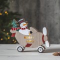 Drewniana świąteczna dekoracja: bałwanek w samolocie z choinką, napis „MERRY X-MAS” na boku, ustawiona na jasnym blacie, w tle rozmyta choinka z lampkami, tworząca radosny, zimowy nastrój.