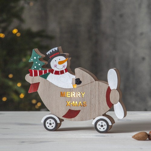 Drewniana świąteczna dekoracja: bałwanek w samolocie z choinką, napis „MERRY X-MAS” na boku, ustawiona na jasnym blacie, w tle rozmyta choinka z lampkami, tworząca radosny, zimowy nastrój.