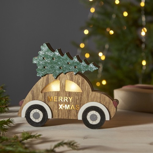 Drewniana podświetlana dekoracja świąteczna w kształcie auta z choinką na dachu i napisem MERRY X-MAS, z ciepłym światłem LED w oknach, ustawiona na stole na tle rozświetlonej choinki.