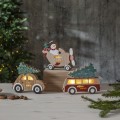 Drewniane dekoracje świąteczne LED MERRY X-MAS – samolot z uśmiechniętym bałwankiem, auto z choinką i bus z drzewkiem, ustawione na drewnianym blacie przy choince, tworzą ciepły, bożonarodzeniowy klimat.