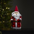 Świąteczna figurka LED Mikołaj w czerwonym stroju i czapce, z długą brodą i ciepłym podświetleniem, stojąca na komodzie na tle choinki i wprowadzająca przytulny klimat świąt.