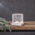 Dekoracja świetlna w formie przezroczystego sześcianu z drobinkami brokatu osadzona na białej podstawie stojąca na drewnianej półce obok gałązki świerku na tle szarej ściany.