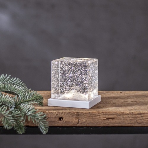 Dekoracja świetlna w formie przezroczystego sześcianu z drobinkami brokatu osadzona na białej podstawie stojąca na drewnianej półce obok gałązki świerku na tle szarej ściany.